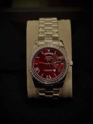 NIBOSI klocka med röd urtavla - Stilren klocka från NIBOSI med metallarmband i silver och en snygg röd urtavla. Klockan har dag- och datumvisning samt romerska siffror på urtavlan. Perfekt accessoar för dig som vill sticka ut med en klassisk men modern look.