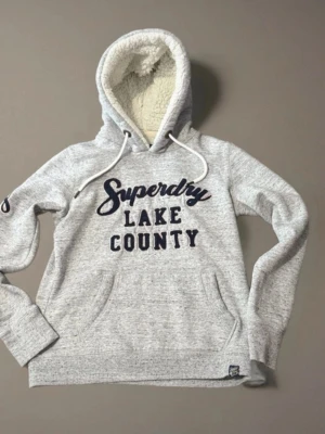 Grå Superdry hoodie med teddyfoder - Mysig ljusgrå hoodie från Superdry med broderad text 'Superdry Lake County' på bröstet. Utrustad med stor huva fodrad med vitt teddyfoder och dragsko samt känguruficka framtill. Perfekt för dig som gillar sportig och avslappnad stil.