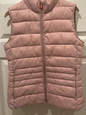 Ljusrosa dunväst med dragkedja - Snygg ljusrosa dunväst med hög krage och horisontella sömmar. Västen har en hel dragkedja framtill och är lätt vadderad för extra värme. 