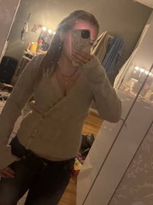 Beige omlottkofta med knappar - Snygg beige omlottkofta med djup v-ringning och dekorativa knappar framtill. Långa ärmar och mjukt stickat material som känns skönt mot huden. Perfekt att styla med jeans eller kjol för en trendig look.