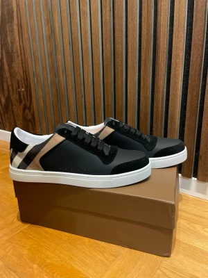 Burberry svarta sneakers med rutmönster - Snygga svarta sneakers från Burberry med klassiskt beige, svart och vitt rutmönster på sidan. Skorna har vita sulor, svarta skosnören och detaljer i mocka. Perfekta för dig som vill ha en stilren och exklusiv look.