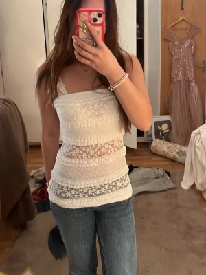 Vit bandeau tubtopp med spetsdetaljer - Säljer en söt vit bandeau tubtopp med genombrutna spetsdetaljer och blommönster. Toppen är elastisk och sitter tight mot kroppen, perfekt till jeans eller kjol. Materialet känns lätt och luftigt, vilket gör den skön att bära under varma dagar.