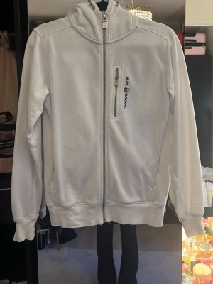 Vit hoodie från Sail Racing - Vit hoodie med dragkedja från Sail Racing. Har en huva, bröstficka med dragkedja och logga på vänster sida. Tillverkad i mjukt material, perfekt för en avslappnad stil. Långa ärmar och ribbade muddar vid ärmslut och nederkant.