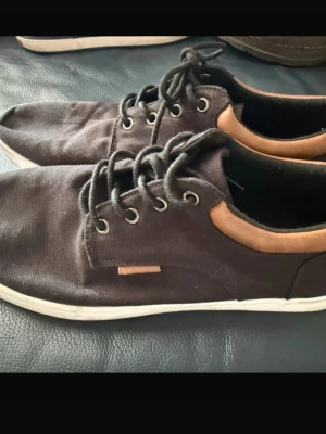 Svarta sneakers från Pier One - Säljer ett par svarta sneakers från Pier One med snygga bruna detaljer runt hälen. Skorna har snörning, vit platt sula och är tillverkade i textil med inslag av skinnimitation. Perfekta för dig som gillar stilrena och enkla skor.