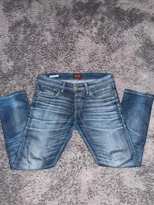 Blåa jeans från Jack & Jones  - Säljer ett par blåa jeans från Jack & Jones med klassisk femficksdesign och raka ben. Jeansen har snygga slitningar och en läderpatch med logga bak i midjan. Tillverkade i bomull för en skön och avslappnad känsla.
