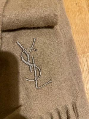 Beige halsduk från Yves Saint Laurent - Snygg beige halsduk från Yves Saint Laurent i mjuk ullmix. Halsduken har klassisk YSL-logga broderad i vitt och fransar i kanterna. Perfekt accessoar för att lyfta din outfit med en lyxig touch.