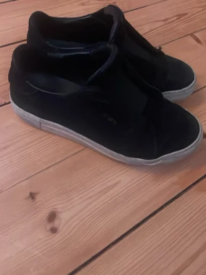Svarta slip-on sneakers från Axel Arigato - Säljer ett par svarta slip-on sneakers från Axel Arigato. Skorna har en stilren design med ovandel i mocka och elastiska paneler för enkel påtagning. Vit sula i gummi ger en snygg kontrast till det svarta. Perfekta för en clean och avslappnad look.