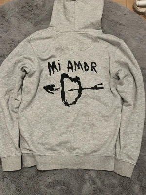 Grå hoodie med tryck 'Mi Amor' - Snygg grå hoodie med svart tryck på ryggen där det står 'Mi Amor' och en pil genom ett hjärta. Klassisk känguruficka framtill och dragsko i huvan. Perfekt för dig som gillar streetwear och unika prints.