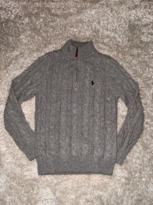 Grå kabelstickad tröja Polo Ralph Lauren - Snygg grå kabelstickad tröja från Polo Ralph Lauren med halv dragkedja och hög krage. Tröjan har det klassiska broderade logotypen på bröstet och ribbade muddar vid ärmslut och nederkant. Perfekt för lager på lager och riktigt stilren.