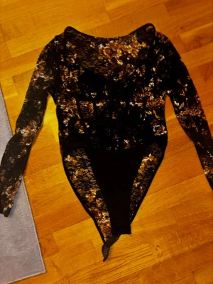 Svart spetsbody med blommönster - Snygg svart body med långa ärmar i transparent spets och blommönster i guld och beige toner. Kroppsnära passform och djup urringning. Perfekt för dig som vill sticka ut med coola detaljer och elegant look.