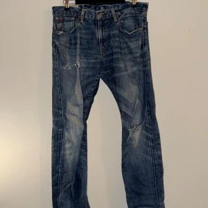 Ralph Lauren  blå jeans herr - Klassiska blå jeans från Ralph Lauren  med rak passform och slitna detaljer på framsidan. Jeansen har fem fickor, knappgylf och tydliga tvättade partier för en vintage look. Tillverkade i slitstark denim, perfekt för en avslappnad stil.