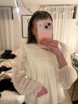 Vit blus med volangdetaljer - Supersöt vit blus med transparenta långärmar, volangdetaljer och diskret strukturmönster. Blusen har rynk vid bröstet och små volanger vid hals och ärmslut. Knapp i nacken, från Gina Young så den är i storlek 170