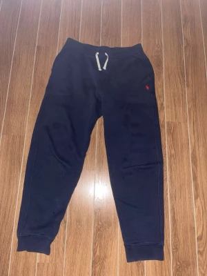 Mörkblå mjukisbyxor från Polo Ralph Lauren - Snygga mörkblå mjukisbyxor från Polo Ralph Lauren med vit dragsko i midjan och den klassiska röda loggan broderad på vänster sida. Byxorna har muddar vid bensluten och är gjorda i mjukt bomullsmaterial. Perfekta för chill dagar eller när du vill vara bekväm. Storlek L (14-16). Bara hör av er vid funderingar!😃