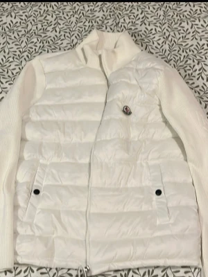 Vit dunväst från Moncler - Snygg vit dunväst från Moncler med quiltad design och dragkedja. Västen har en klassisk look och är fodrad med mjukt material. Perfekt att slänga över en hoodie eller tröja för extra värme. Märkeslogga på insidan och coola detaljer.