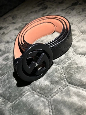 Svart skärp med GG-spänne från Gucci - Snyggt svart bälte från Gucci med klassiskt GG-spänne i svart metall. Bältet är gjort i skinn och har diskret präglat mönster längs remmen. Insidan är beige och ger en lyxig kontrast. Perfekt accessoar för att lyfta din outfit.