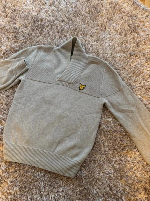 Grå stickad tröja från Lyle & Scott - Snygg grå stickad tröja från Lyle & Scott med bred sjalkrage och ribbade muddar. Tröjan har det klassiska gula örnloggan broderad på bröstet och är tillverkad i mjukt material. Perfekt för lager-på-lager och stilren look.