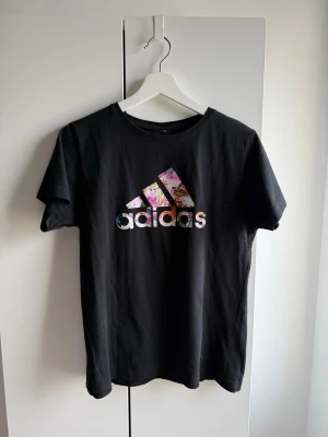 Svart Adidas t-shirt med blommigt tryck - •Snygg svart t-shirt från Adidas med klassiskt logotryck framtill i färgglatt blommigt mönster.  •Mycket bra skick. Sparsamt använd. •Storlek M, alla etiketter är bortklippta.  Djurfritt och rökfritt hem