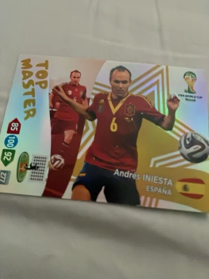 Fotbollssamlarkort Top Master Iniesta - Samlarkort med holografisk yta från FIFA World Cup Brasil, föreställande Andres Iniesta i Spaniens landslagströja. Kortet har texten 'Top Master' och visar flagga, statistik och turneringslogga. Perfekt för fotbollsentusiasten.