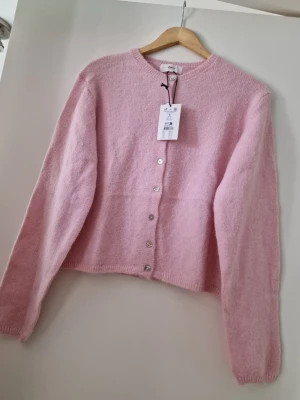 Ljusrosa kofta från Gina Tricot - Supersöt ljusrosa kofta från Gina Tricot med rund halsringning och silvriga knappar framtill. Koftan är stickad i ett mjukt material och har långa ärmar samt ribbade muddar. Perfekt för att addera en feminin touch till din outfit.