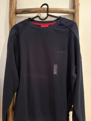 Mörkblå sweatshirt från Hugo boss - Snygg mörkblå sweatshirt från Hugo med diskret logga på bröstet och ton-i-ton Hugo-detaljer längs ärmarna. Tröjan har rund halsringning och är tillverkad i mjukt bomullsmaterial. Perfekt för en clean och stilren look.