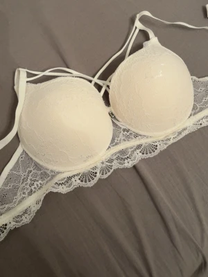 Vit spets-bh med vadderade kupor - Snygg vit bh med vadderade kupor och dekorativa spetsdetaljer. Bh:n har tunna axelband och extra band över bysten för en trendig look. Spetsen ger en romantisk känsla och bh:n har en bred spetskant nedtill.