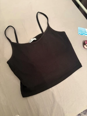 Svart ribbad linnetopp new yorker - Svart ribbad linnetopp från new yorker med smala axelband och croppad passform. Perfekt att styla med jeans eller kjol för en trendig look. Enkel och stilren design som passar till många outfits.