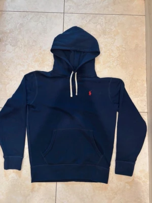 Mörkblå hoodie från Polo Ralph Lauren - Mörkblå hoodie från Polo Ralph Lauren med klassisk huva och vita dragsnören. Har en känguruficka framtill och den ikoniska röda logotypen broderad på bröstet. Tillverkad i mjuk bomullsblandning som känns skön mot huden. Perfekt för en avslappnad och stilren look.