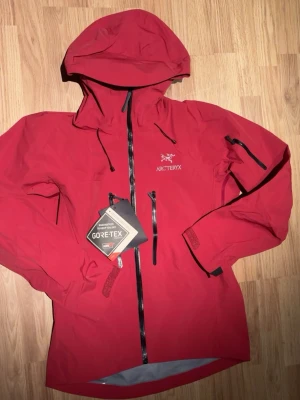 Röd Arc'teryx regnjacka Gore-Tex - Snygg röd regnjacka från Arc'teryx med vattentätt Gore-Tex-material. Jackan har huva, hel dragkedja framtill, justerbara muddar och flera dragkedjeförsedda fickor. Perfekt för blöta dagar och outdoor-äventyr. Logga på bröstet och detaljer i svart. Perfekt för skidåkning, hiker, klättring och allt liknande eller som vanlig regnjacka. Jackan ser ljusare ut på bild än vad den är i verkligheten.