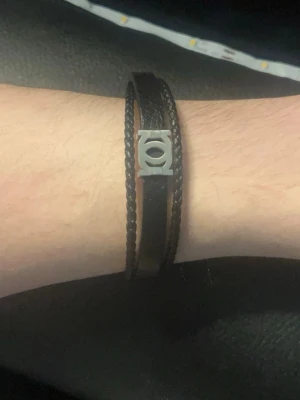 Svart läderarmband med metallspänne - Snyggt svart armband i flätat läder med ett stilrent metallspänne i silverfärg. Armbandet har en enkel och modern design som passar perfekt till en avslappnad eller trendig look. Metallspännet ger en cool detalj och armbandet är lätt att matcha med andra accessoarer.                              Vet inte vilket märke men gissar på police lifestyle.  (Den har ej använts)