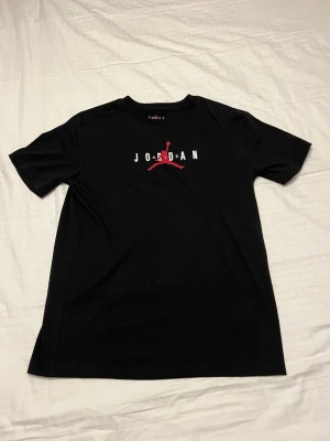 Svart Jordan t-shirt med logga - Snygg svart t-shirt från Jordan med det klassiska Jumpman-trycket i rött och vitt på bröstet. T-shirten har rund hals och korta ärmar, perfekt för en sportig och avslappnad stil. Tillverkad i mjuk bomull för skön komfort.