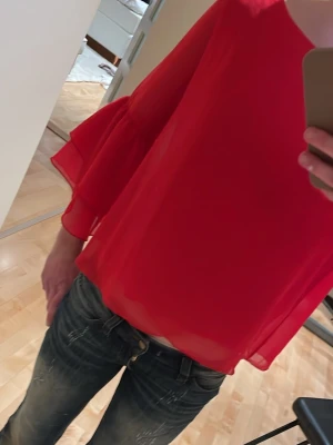 Röd volangblus i transparent tyg - Snygg röd blus i ett lätt och transparent material med vida, volangprydda långärmar. Blusen har en lös passform och rund halsringning. Perfekt för dig som vill sticka ut med en färgstark och trendig look.