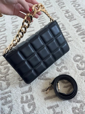 Svart quiltad handväska med guldkedja - Stilren svart handväska i quiltad design med en chunky guldfärgad kedja som handtag. Väskan har en rektangulär form och medföljer en avtagbar axelrem. Perfekt för dig som gillar trendiga accessoarer med lyxig känsla.