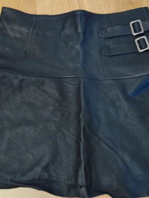 Svartgrå kort kjol med spännen, storlek S - Kjolen är ifrån Hunkydory i storlek S. Snygg svart kort kjol i läder och polyester med två dekorativa spännen i midjan. Kjolen har hög midja och en clean, stilren look som passar till många olika outfits. Perfekt för dig som vill ha en edgy och trendig stil. 