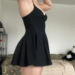 Svart kort klänning med v-ringning - Snygg svart kort klänning med smala axelband och lätt utställd kjol. Klänningen har en liten knapp framtill och en feminin, figurnära passform upptill. Klänningen är i ett glatt och mycket stretchigt material och har även inbyggda shorts. Klänningen saknar både märke och storlek, men skulle gissa på strl S. Knappen är något hängig men går att enkelt sy fast bättre