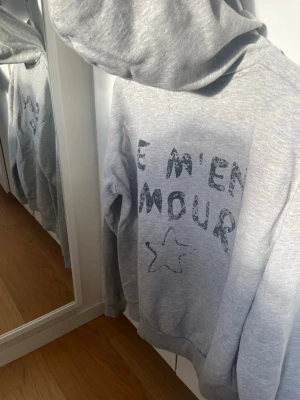 Grå hoodie med tryck och stjärna - Grå hoodie med svart texttryck 'JE M'EN AMOUR' och en handritad stjärna på bröstet. Klassisk passform med huva och ribbade muddar. Perfekt för en avslappnad och trendig look.