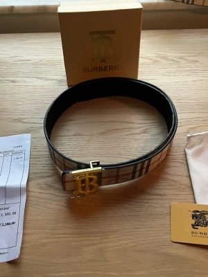 Burberry rutigt bälte med guldfärgat spänne - 105 cm, Snyggt bälte från Burberry i klassiskt beige rutigt mönster med inslag av rött och svart. Bältet har ett stort guldfärgat spänne med Burberrys ikoniska B-logga. Perfekt accessoar för att lyfta din outfit.