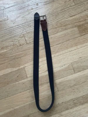 Mörkblått flätat bälte från Saddler - Snyggt mörkblått flätat bälte från Saddler med detaljer i brunt skinn och silverfärgat spänne. Perfekt accessoar för att lyfta din outfit och funkar till både jeans och chinos.