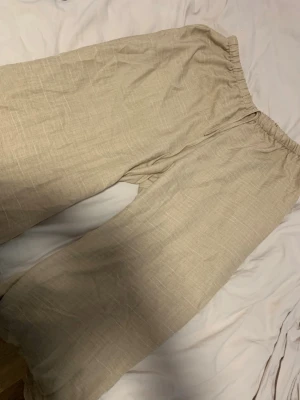 Beiga vida linnebyxor med resår - Säljer ett par beiga linnebyxor med bred passform och resår i midjan. Byxorna är tillverkade i ett luftigt linnematerial och har en avslappnad look som passar perfekt till sommaren. Inga synliga fickor eller detaljer. Märke ej synligt.