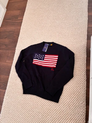 Mörkblå stickad tröja Polo Ralph Lauren - Snygg mörkblå stickad tröja från Polo Ralph Lauren med amerikansk flagga i rött, vitt och blått på bröstet. Rund halsringning och ribbade muddar vid ärmslut och nederkant. RL-broderi i rött nedanför flaggan. Perfekt för dig som gillar klassisk preppy stil.