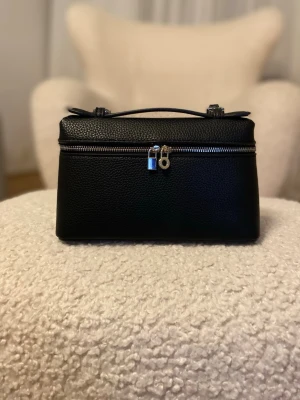 Svart handväska med silverdetaljer - Stilren svart handväska i strukturerat läderimitation med silverfärgade detaljer och dragkedja. Väskan har ett topphandtag och dekorativa låsdetaljer framtill. Perfekt för dig som gillar minimalistisk och modern design.