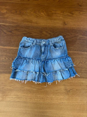 Blå volangkjol i denim XS Gina tricot  - Säljer en blå jeanskjol med volanger och råa kanter. Kjolen har två fickor fram, knapp och dragkedja samt bälteshällor. Perfekt för dig som gillar en avslappnad och trendig stil.