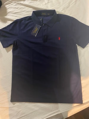 Marinblå polotopp från Polo Ralph Lauren - Säljer en klassisk marinblå polotopp från Polo Ralph Lauren med korta ärmar och krage. Toppens ikoniska röda logga är broderad på bröstet och den har knappar framtill. Perfekt för dig som gillar stilrena och tidlösa plagg.