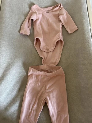 Ljusrosa body och byxa set - Säljer ett set med ljusrosa body och matchande byxa. Bodyn har långa ärmar, rund hals och tryckknappar i grenen för enkel påklädning. Byxorna är mjuka och har resår i midjan. Perfekt för en bekväm och stilren look.