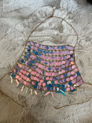 Festivaltopp holografisk  - Unik topp med stora paljetter i holografisk färg. kedjedetaljer i guld och halterneck-modell. Toppen har glittrande effekt och är perfekt för att sticka ut. Justerbara band i sidorna och dekorativa hängande paljetter längst ner. Perfekt till sommarfesten, festival, rave och party 😍☀️