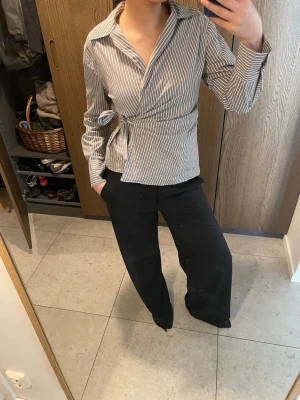 Randig omlottblus med knyt i midjan - Snygg randig omlottblus i vitt och grått med knyt i midjan och klassisk krage. Skjortan har långa ärmar och en figurnära passform som ger en stilren look. Perfekt för dig som gillar trendiga detaljer och vill ha något unikt i garderoben.