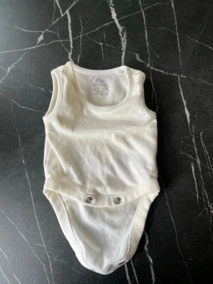 Vit ärmlös body från NEXT baby - Klassisk vit body från NEXT baby i mjuk bomull. Ärmlös modell med rund halsringning och tryckknappar i grenen för enkel påklädning. Perfekt basplagg till garderoben och lätt att matcha med andra plagg.