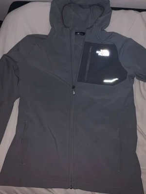 Grå vindjacka från The North Face - Snygg grå vindjacka med huva från The North Face. Jackan har hel dragkedja, bröstficka med dragkedja och reflekterande logga. Tillverkad i ett lätt och slitstarkt material, perfekt för blåsiga dagar. Två sidofickor med knappstängning och meshfoder för extra ventilation.
