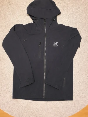 RevolutionRace Svart Hiball Jacka L - Snygg svart vindjacka från RevolutionRace med hel dragkedja, justerbar huva och två bröstfickor med dragkedja. Jackan har minimalistisk design, diskret logga på bröstet och är tillverkad i ett lätt, vindtätt material. Perfekt för blåsiga dagar och enkel att matcha.