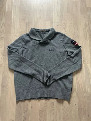 Grå tröja med sjalkrage från woolrich - Mysig grå tröja från woolrich med sjalkrage och knappdetalj. Broderad logga på bröstet och amerikansk flagga som patch på ena ärmen. Långärmad modell med ribbade muddar och nederkant. Perfekt för en avslappnad och stilren look.