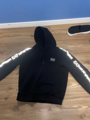 Svart EA7 dress med logga - Svart hoodie från EA7 Emporio Armani med huva. Stora vita logotexter längs båda armarna och benen och EA7-logga på bröstet och på byxorna.Tillverkad i mjukt material som passar perfekt för en avslappnad och sportig stil.